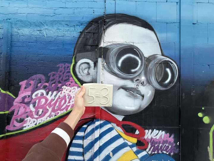 A hand holding a 5=3 plaque in front of a mural, Bremerhaven